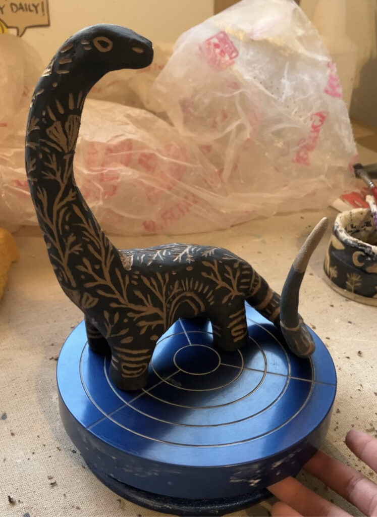 A pre-fired brontosaur sculpture done in a black with sgraffito details.
