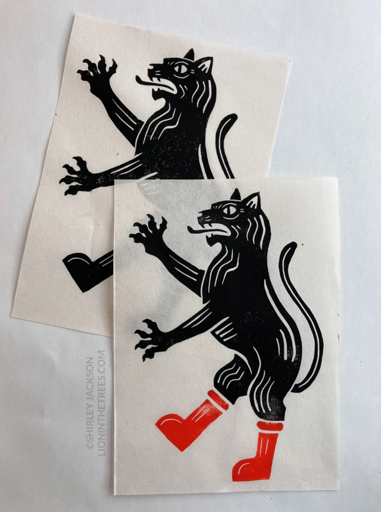 Photo of two relief block prints featuring a heraldic style black cat on white/natural coloured paper. The print on top/in the foreground has red Astro Boy boots and the print behind that one has black boots.