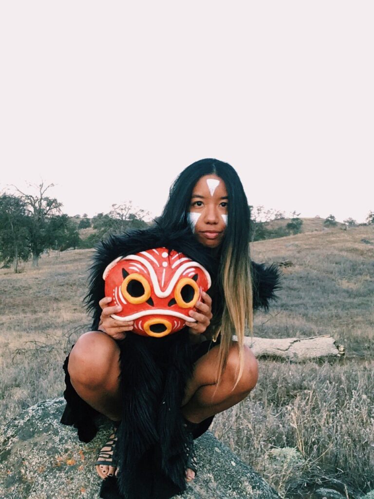 It's me, ya girl, in my DIY San outfit crouching on a rock looking cute holding the mask off, but in front of me showing it off. You can see the white upside down triangle markings I've painted on my face.