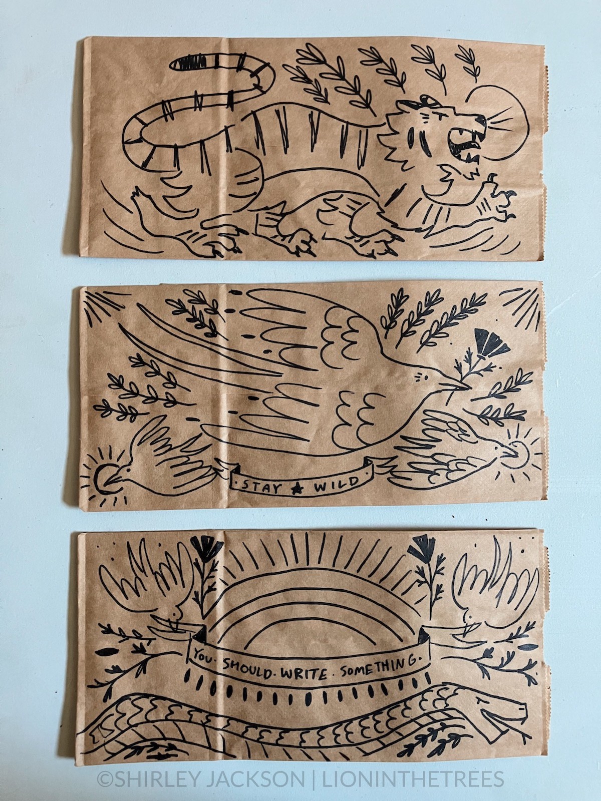 A photo of 3 brown paper lunch bags with a variety of doodles drawn with a sharpie on them. This variety features subject matter ranging from a tiger, a snake, and several barn swallows with lettering and plant-like motifs.
