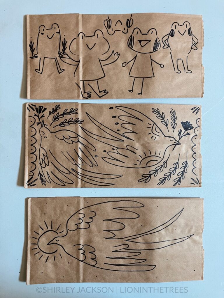 A photo of 3 brown paper lunch bags with a variety of doodles drawn with a sharpie on them. This variety features subject matter ranging from an anthropomorphic frog and more barn swallow motifs.
