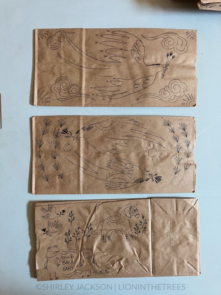 A photo of 3 brown paper lunch bags with a variety of doodles drawn with a pen on them. This variety features subject matter ranging from barn swallows, Kermit the Frog, and a dala horse