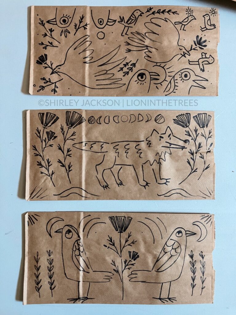 A photo of 3 brown paper lunch bags with a variety of doodles drawn with a sharpie on them. This variety features subject matter ranging from different birds, wolves, and California Poppy motifs.
