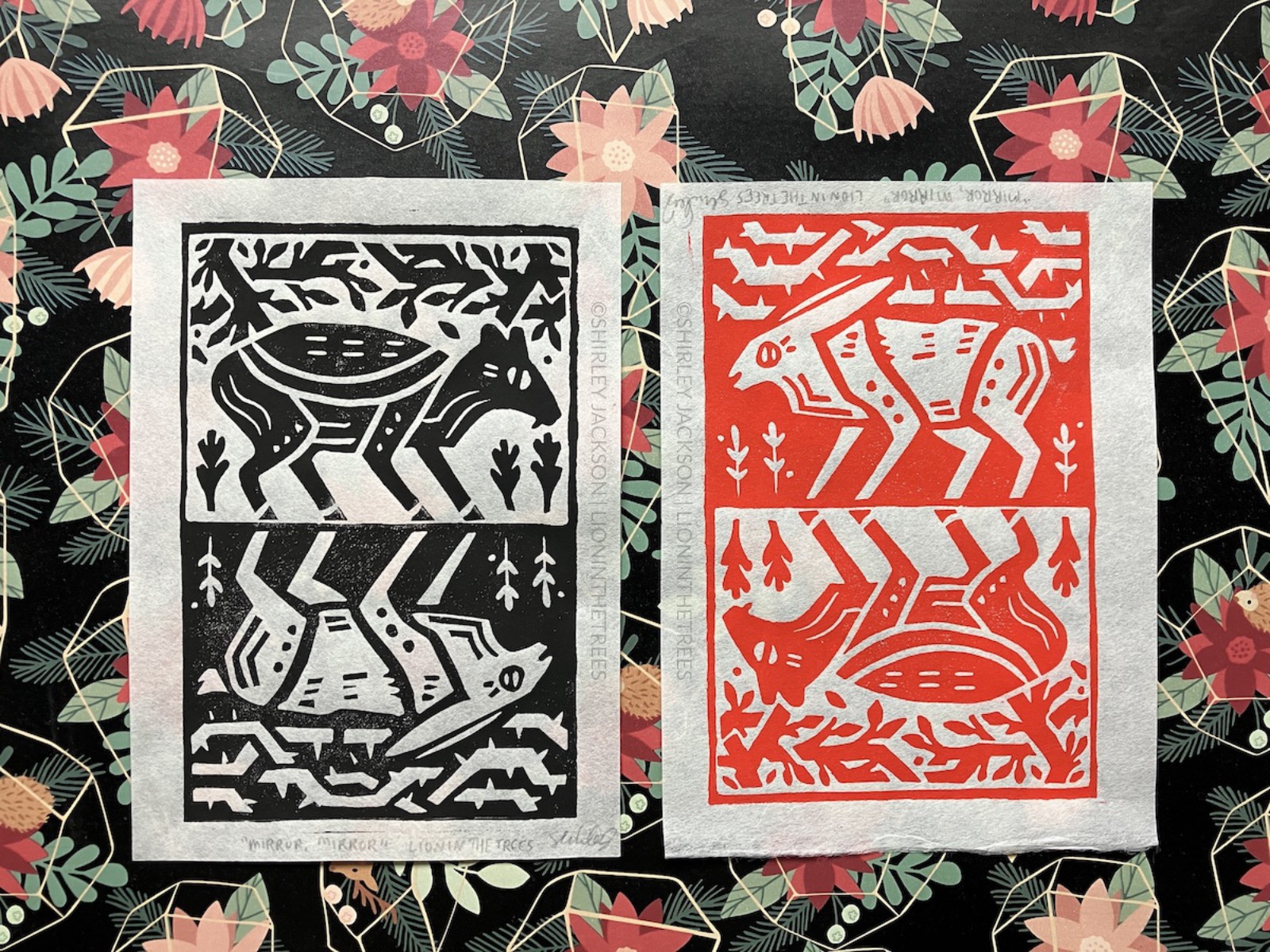 A photo showing both colour variants of the Sang Kancil print. There is a black one on the left and a red one on the right. The print features two animals mirroring one another. At the top of the print is Sang Kancil, a small Mouse-Deer. They are printed in black against white with tree branches and leaves above them. Mirror reflected below Sang Kancil is Br'er Rabbit. Br'er Rabbit is printed white against black and features barbed branches of a briar bush instead of tree branches and leaves.