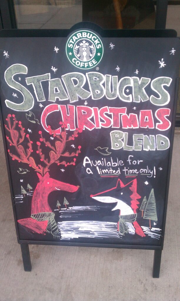 Photo of a chalkboard sign that features their Christmas Fox and Christmas Reindeer mascots with the words "Starbucks Christmas Blend" and "Avaiilable for a limited time only!" handdrawn on the chalkboard.