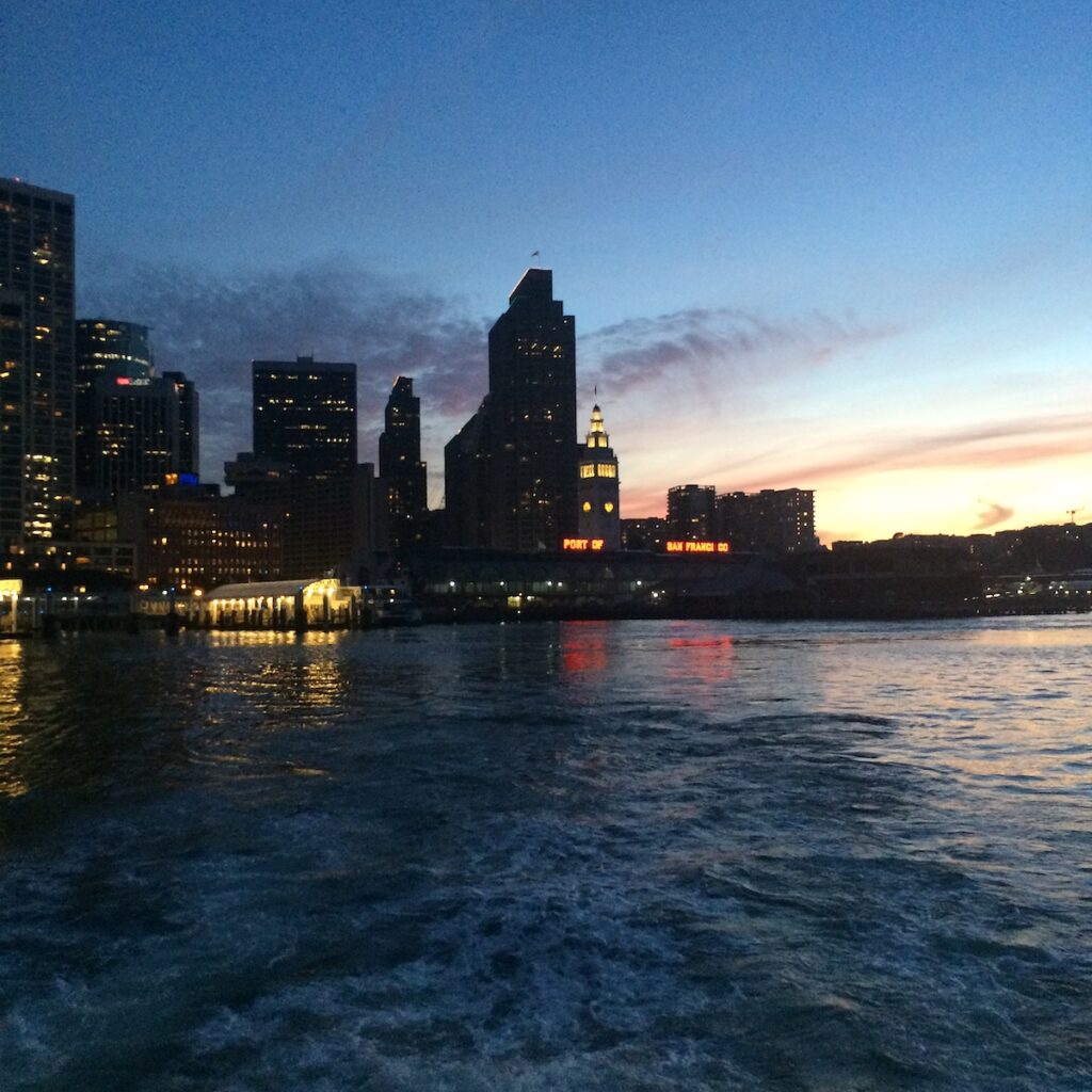 Photo taken from the back of a shuttle boat leaving San Francisco. The photo was taken at twilight so the sun is starting to set and the buildings are silhouetted and their lights are glowing against the darkening sky.