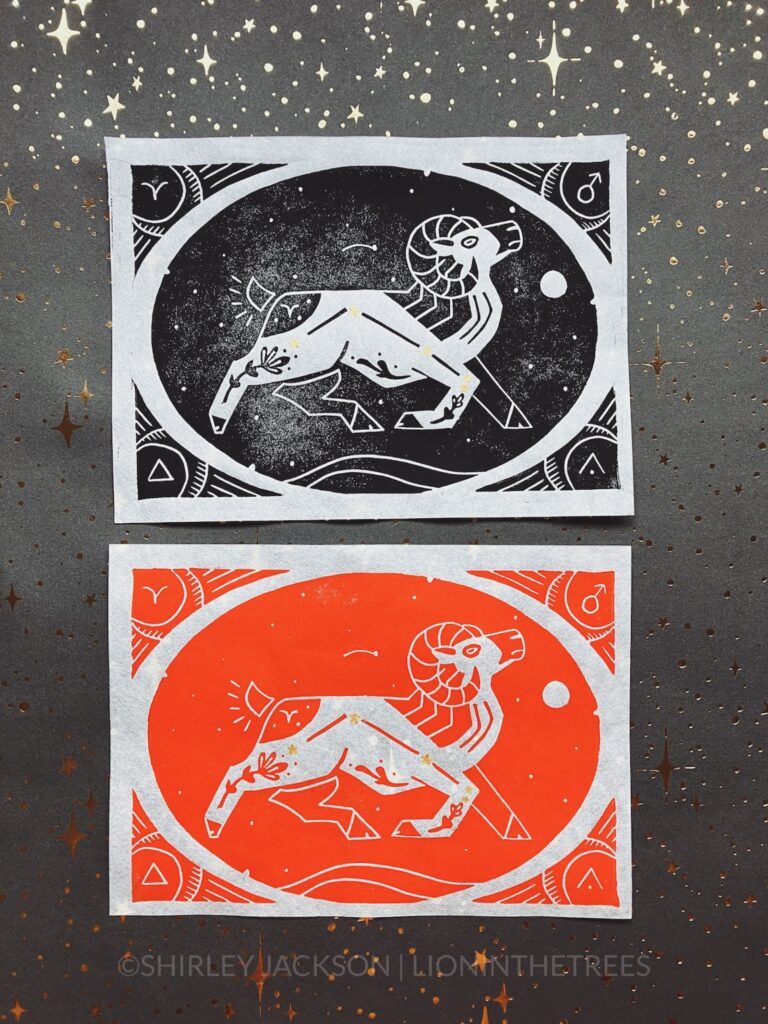 Photo of an all black and an all red block print featuring the astrological sign of Aries as a Bighorn Sheep with the constellation hand-painted with gold stars on the sheep’s body. They are enclosed within an oval that has stars within it, and a border that has symbolic signs that coordinate with Aries.