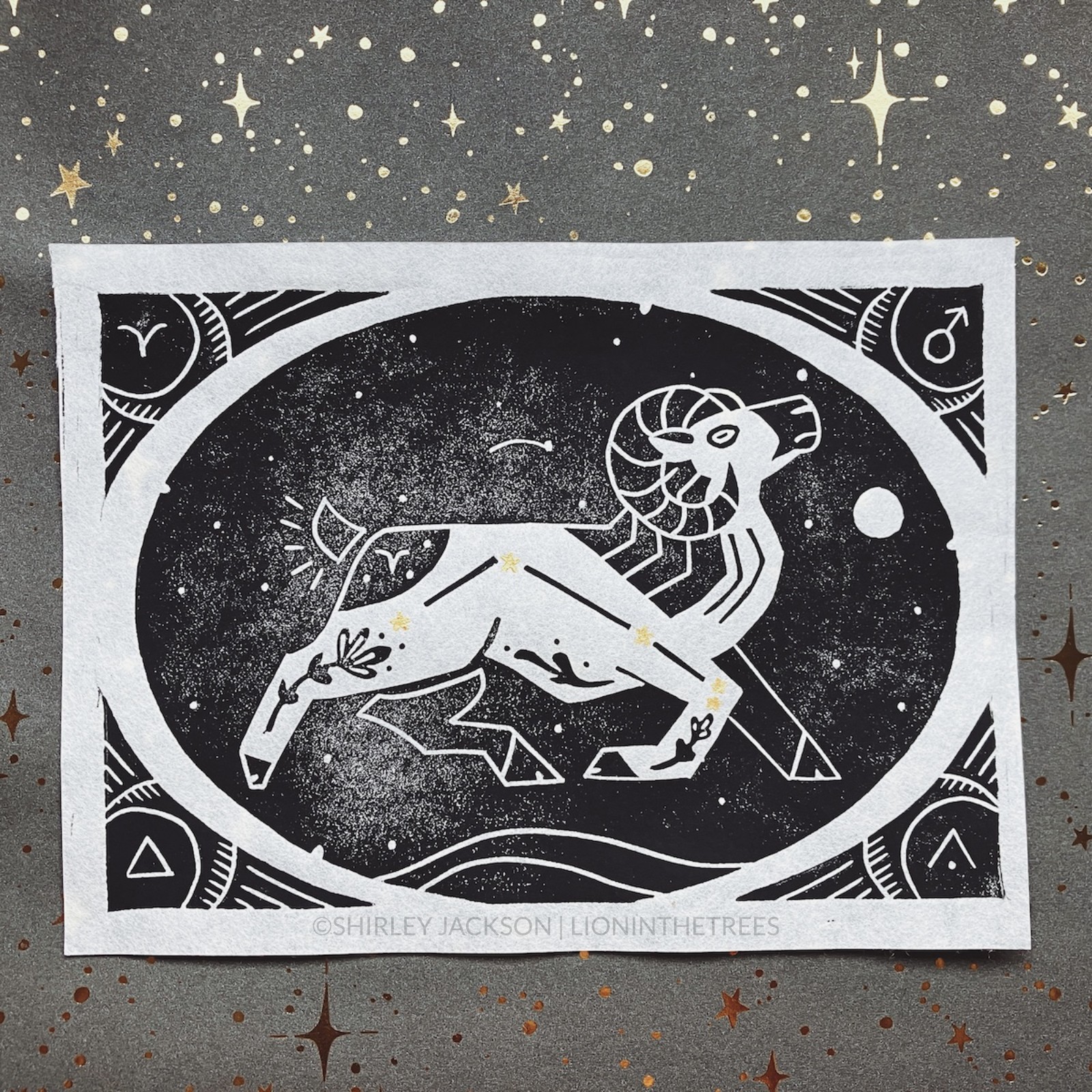 A black block print featuring the astrological sign of Aries as a Bighorn Sheep with the constellation hand-painted with gold stars on the sheep’s body. They are enclosed within an oval that has stars within it, and a border that has symbolic signs that coordinate with Aries.