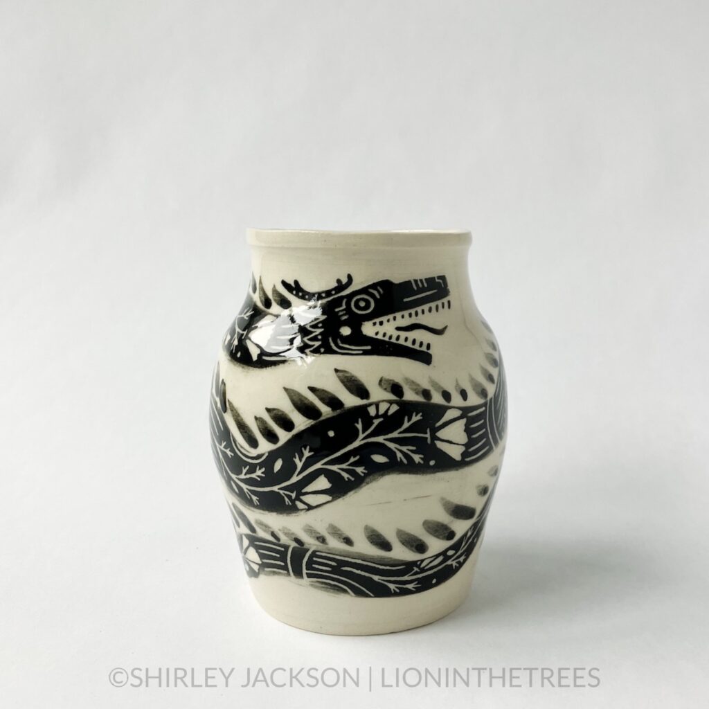A dragon vase featuring an experimental dragon motif done in black underglaze painted directly onto the creamy white body of the clay. This photo shows a side of the jar showing the head of the dragon and parts of it's body as it wraps around the circumference of the vase.