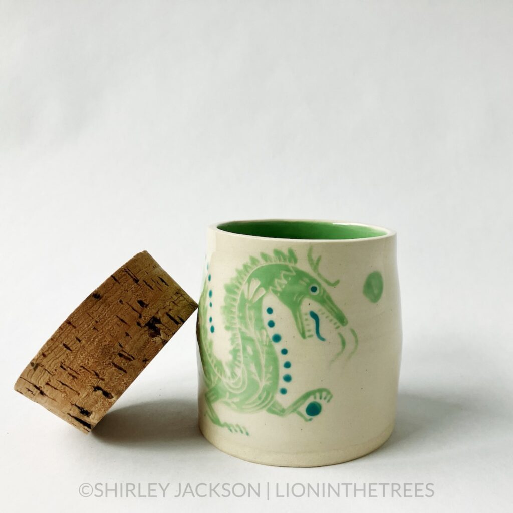 Dragon memory jar with a cork lid featuring an experimental sgraffito dragon motif. This one was done with pistachio green and turquoise underglaze directly painted onto the creamy coloured clay body of the jar. This photo features the cork lid removed and off to the side showing off the green interior.