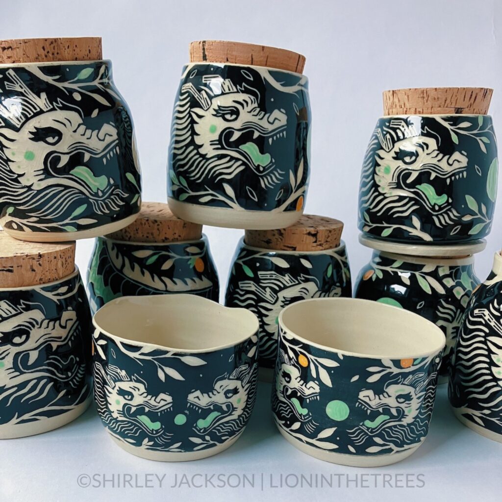 Group photo showing eight dragon memory jars and the small bowl and planter. Each design features the same dragon sgraffito motif done in a black and pistachio green underglaze. Some feature oranges within the design as well.