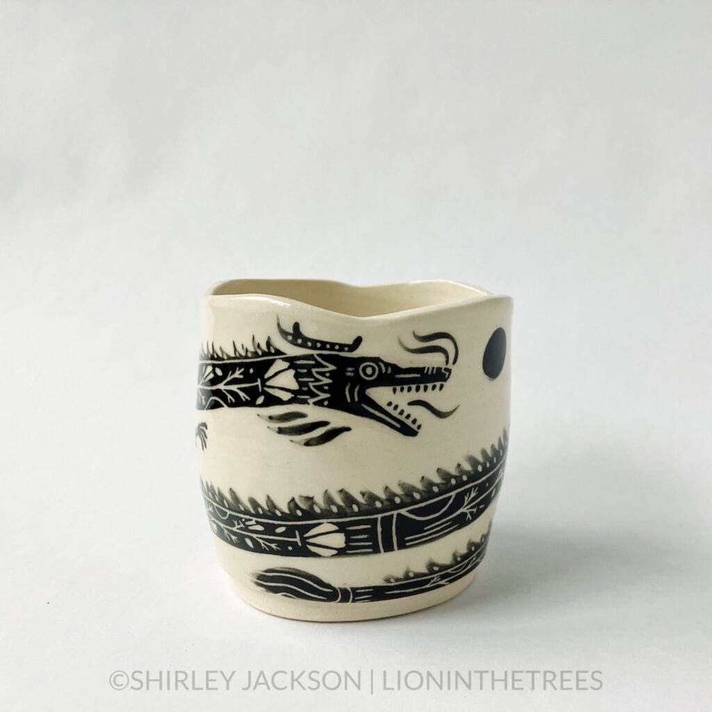 A dragon planter featuring an experimental dragon motif done in black underglaze painted directly onto the creamy white body of the clay. This photo shows a side of the jar showing the head of the dragon and parts of it's body as it wraps around the circumference of the planter.