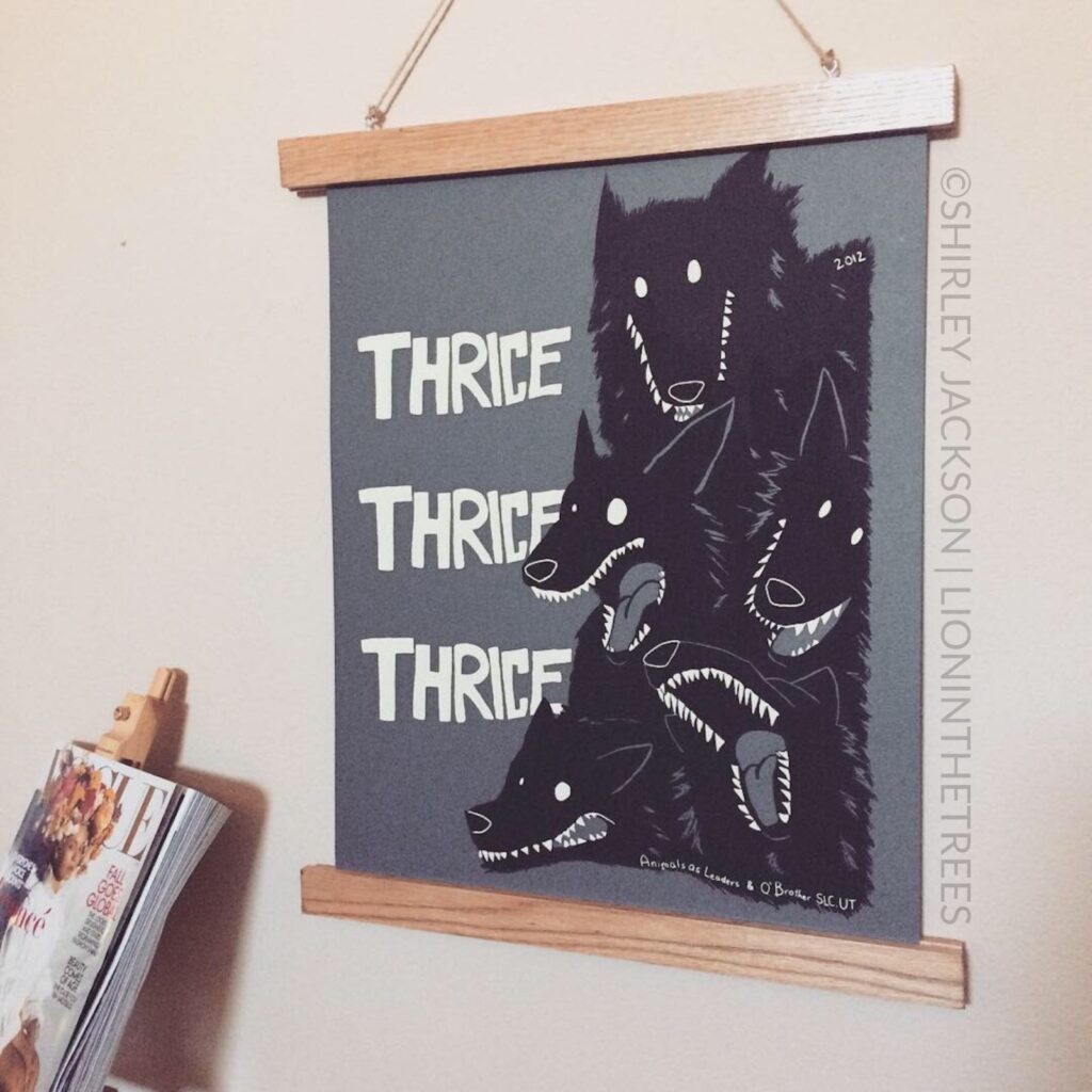 Photo of my Thrice gig poster I made in college hanging up on a wall. It features the word "Thrice" three times, and a pack of black wolves to the right side of the typography printed on a grey paper.