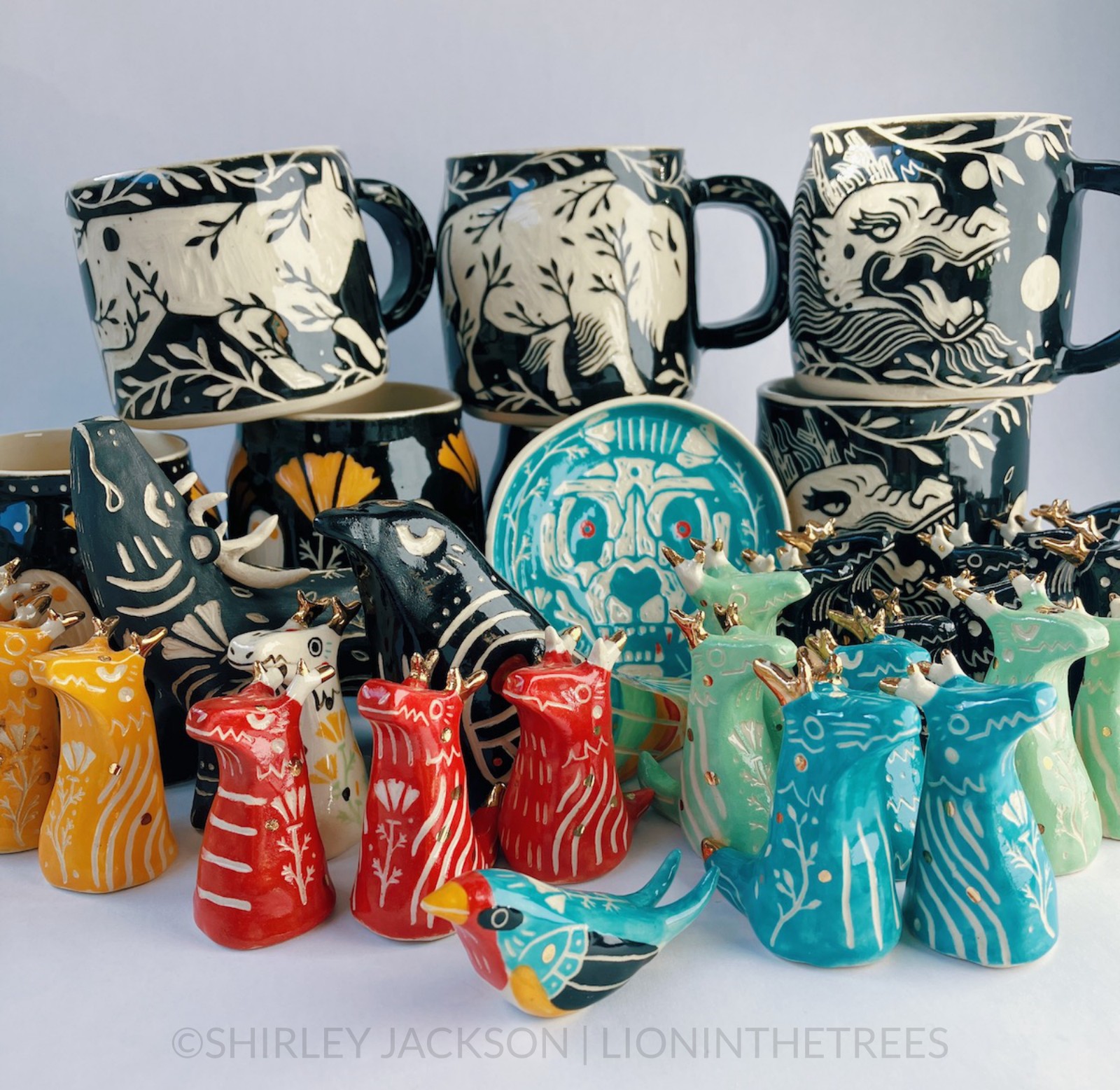 A colourful collection of pottery featuring little dragon totems, a small barn swallow totem, larger animal totems in the back, and further in the back are mugs/vessels stacked on top of each other.