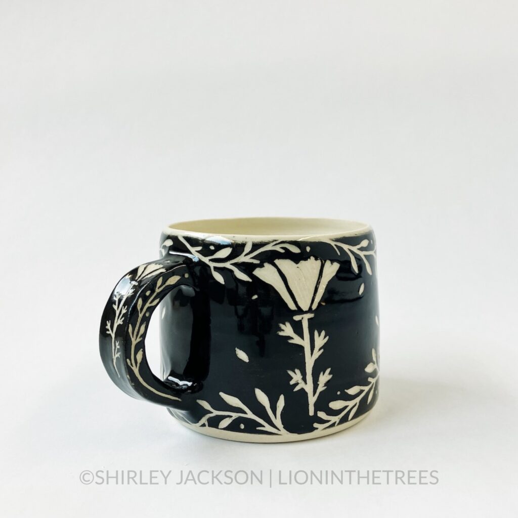 Ceramic black sgraffito mug featuring my Running Wolf motif. This mug also features a floral border above and below the wolf motif. This shows the back that features a California Poppy motif.