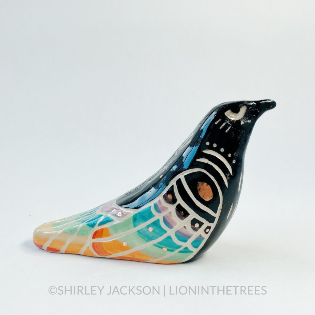 A ceramic sculpture of a crow. The bird has been done in black underglaze, and it’s wings/back feathers have been painted to mimic a rainbow with underglaze, and pops of gold glaze as well. There are sgraffito markings carved around it’s body including a California Poppy on it’s back. Right side view.