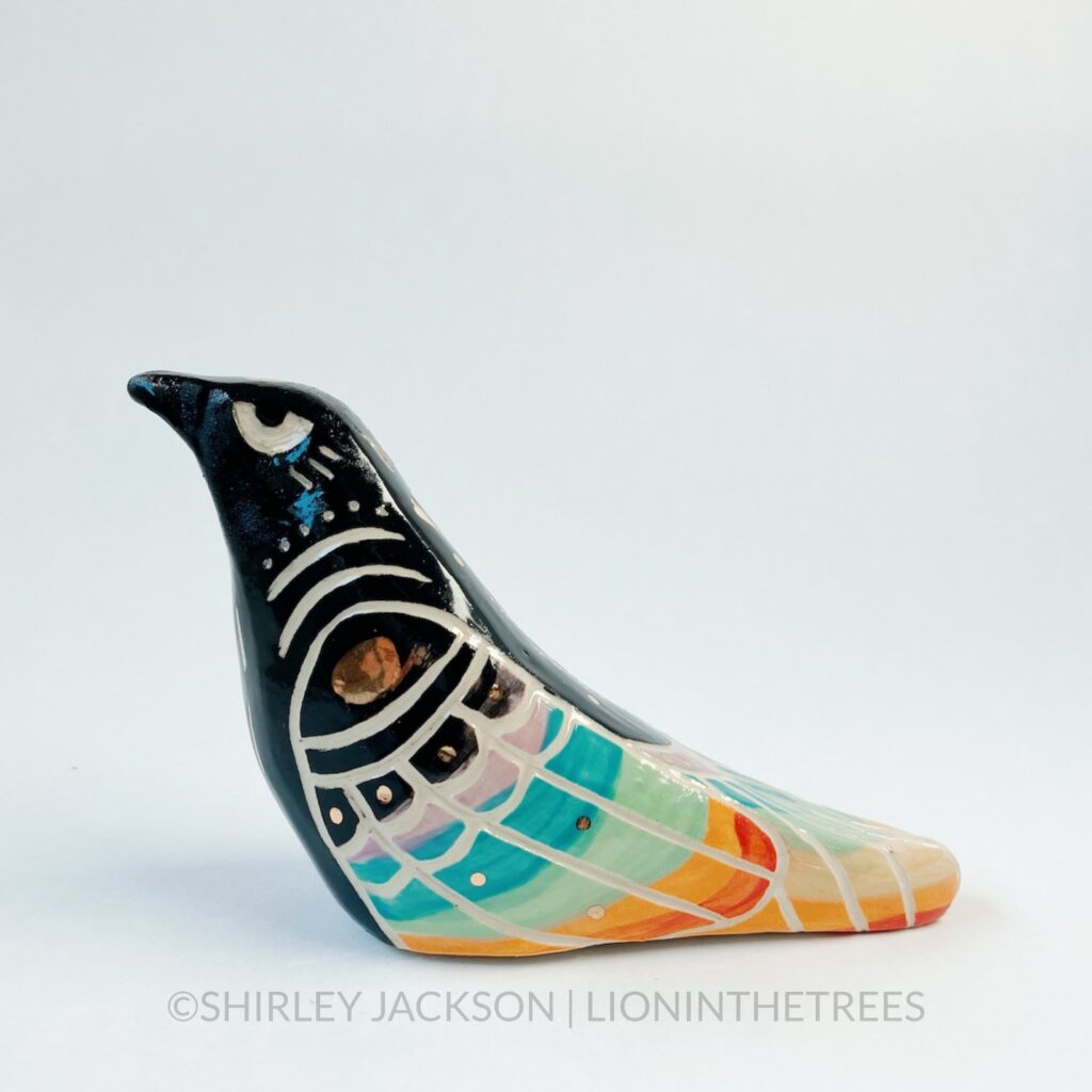 A ceramic sculpture of a crow. The bird has been done in black underglaze, and it’s wings/back feathers have been painted to mimic a rainbow with underglaze, and pops of gold glaze as well. There are sgraffito markings carved around it’s body including a California Poppy on it’s back. Left side view.