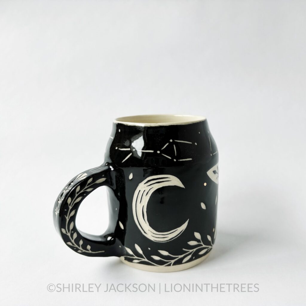 Ceramic black sgraffito mug featuring my Running Wolf motif. This mug also features a floral border above and below the Wolf motif. The back features a carved crescent moon motif.