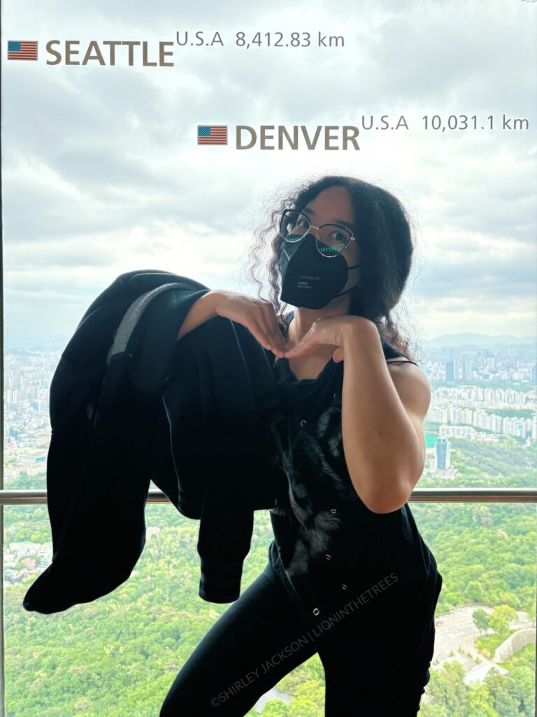 Photo of me posing inside the Seoul tower with the Denver and Seattle location markers on the window above me