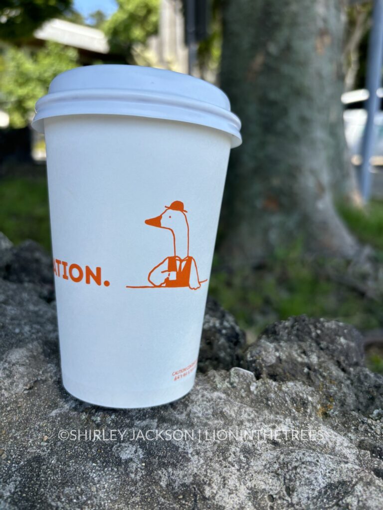 A to-go cup of chai tea with a cartoon goose dressed up in a nice vest and hat on the side of the cup.