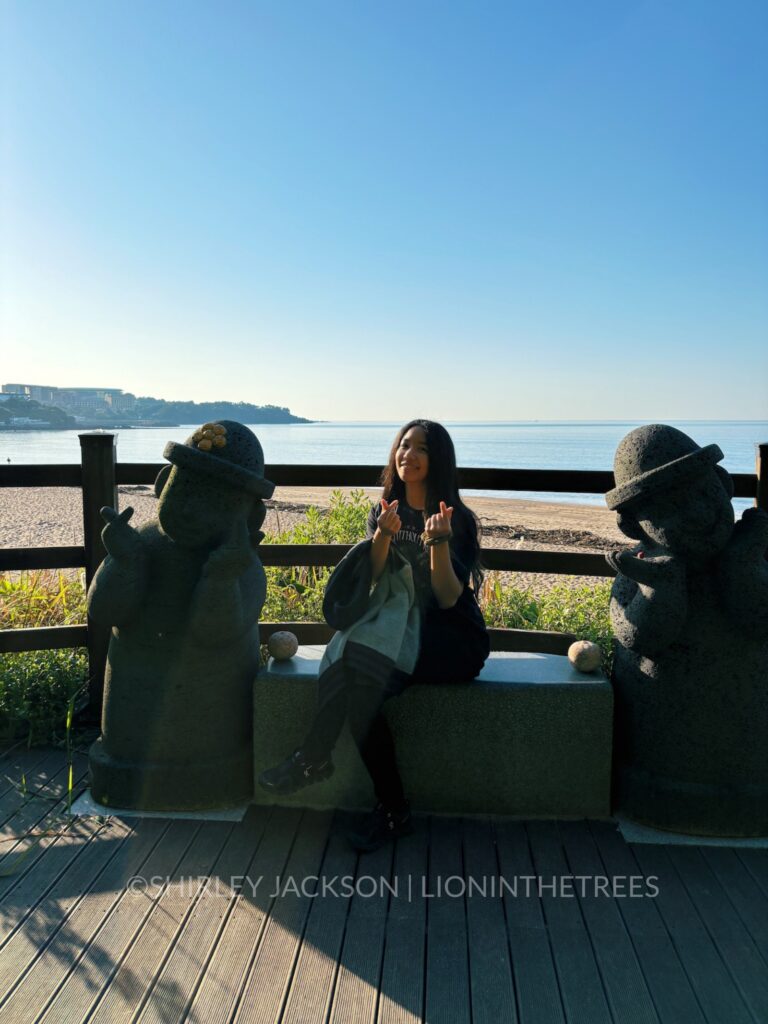 Photo of me posing with two stone Jeju Island dol hareubang in front of the ocean doing the "heart" hand sign.