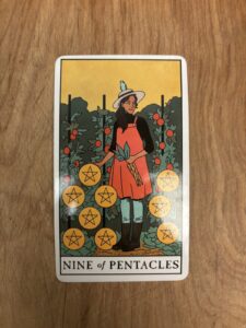 Photo of the 9 of Pentacles card from the Modern Witch tarot deck. It shows an older woman happy and content in her bountiful garden full of ripe tomatoes and pentacles. She's holding a bundle of carrots in one hand, and another is atop a pentacle. She has a little blue bird perched atop her hat as she looks out contently at all that she has.