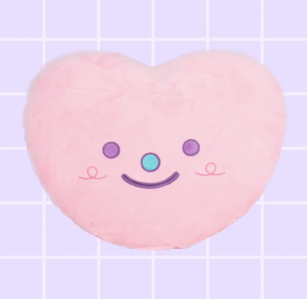 A pink heart shaped pillow with an embroidered smile-y face. Made by Kojo Jojo Studio.