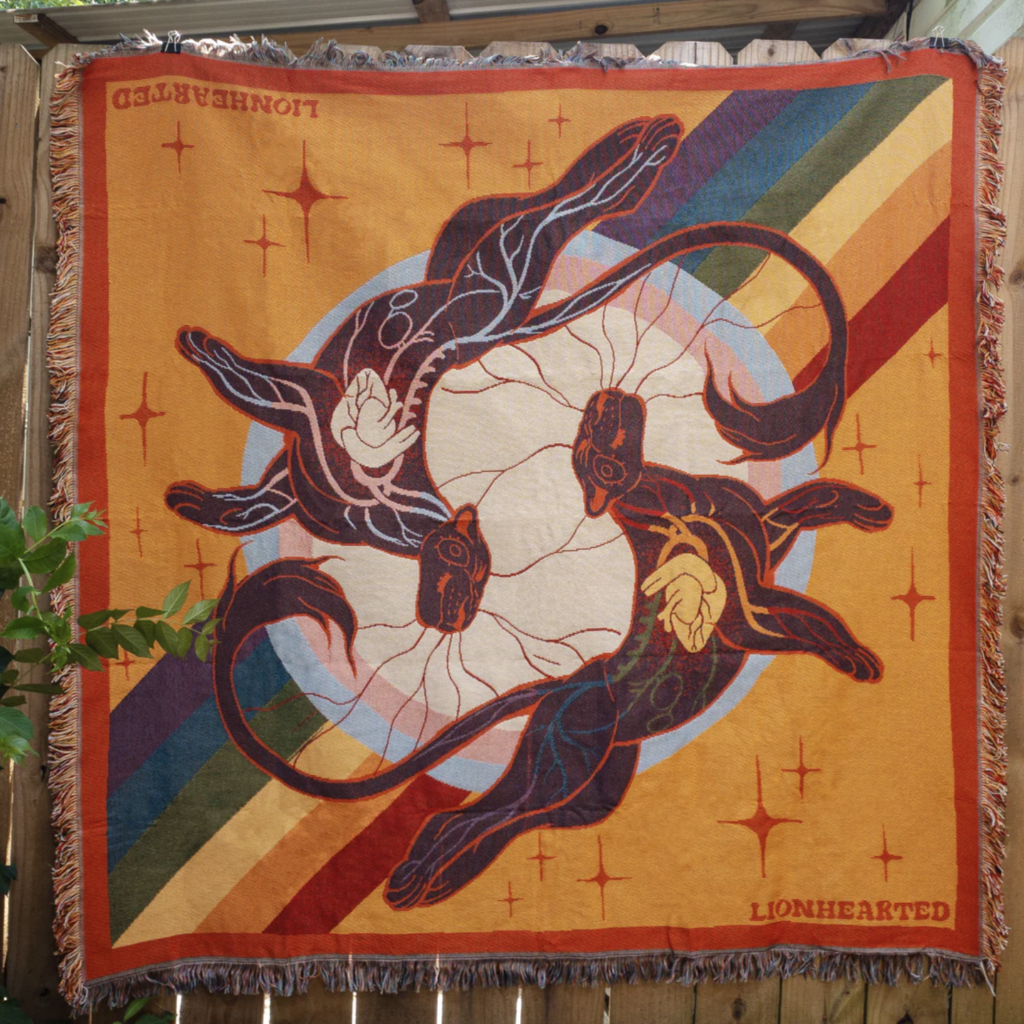 A blanket featuring two lions circling one another that stylistically display their heart and circulatory system. There are the PRIDE rainbow and Trans colours used throughout the piece and within the bodies of the two lions. Made by Aspen Eyes.