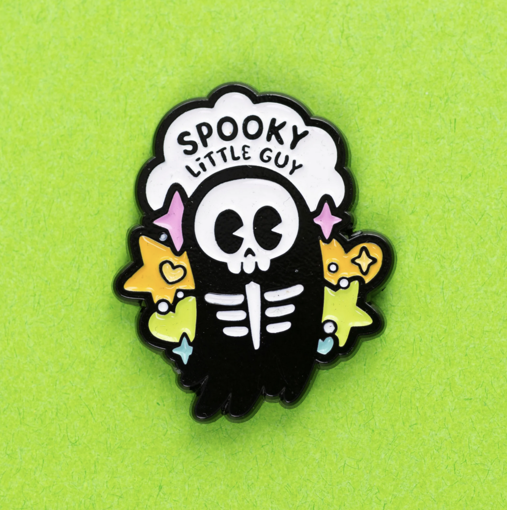 An enamel pin featuring a cute grim reaper figure, pastel coloured stars and hearts behind it with a cloud bubble that says “Spooky little guy”. Made by Pepper Raccoon