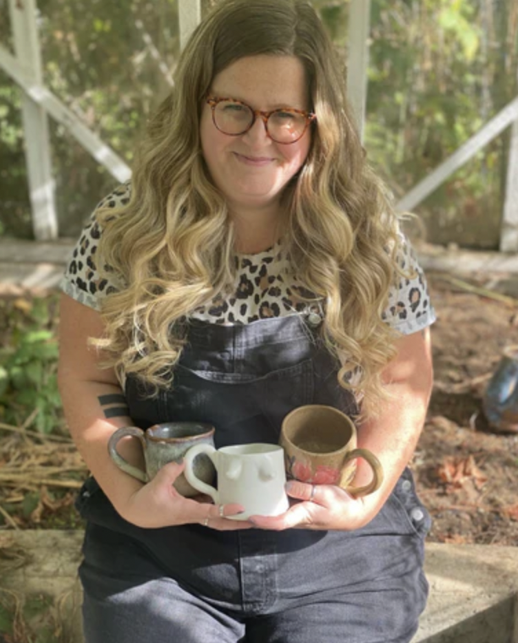 A photo of Nissa Wallace, the artist behind Nissa Arts sitting in a garden holding some of her pottery.