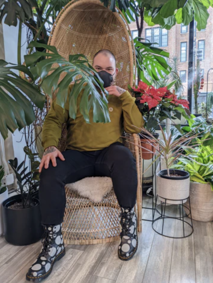 Photo of Leo, the artist behind Chicago Queer Creations seated in this gorgeous room surrounded by plants.