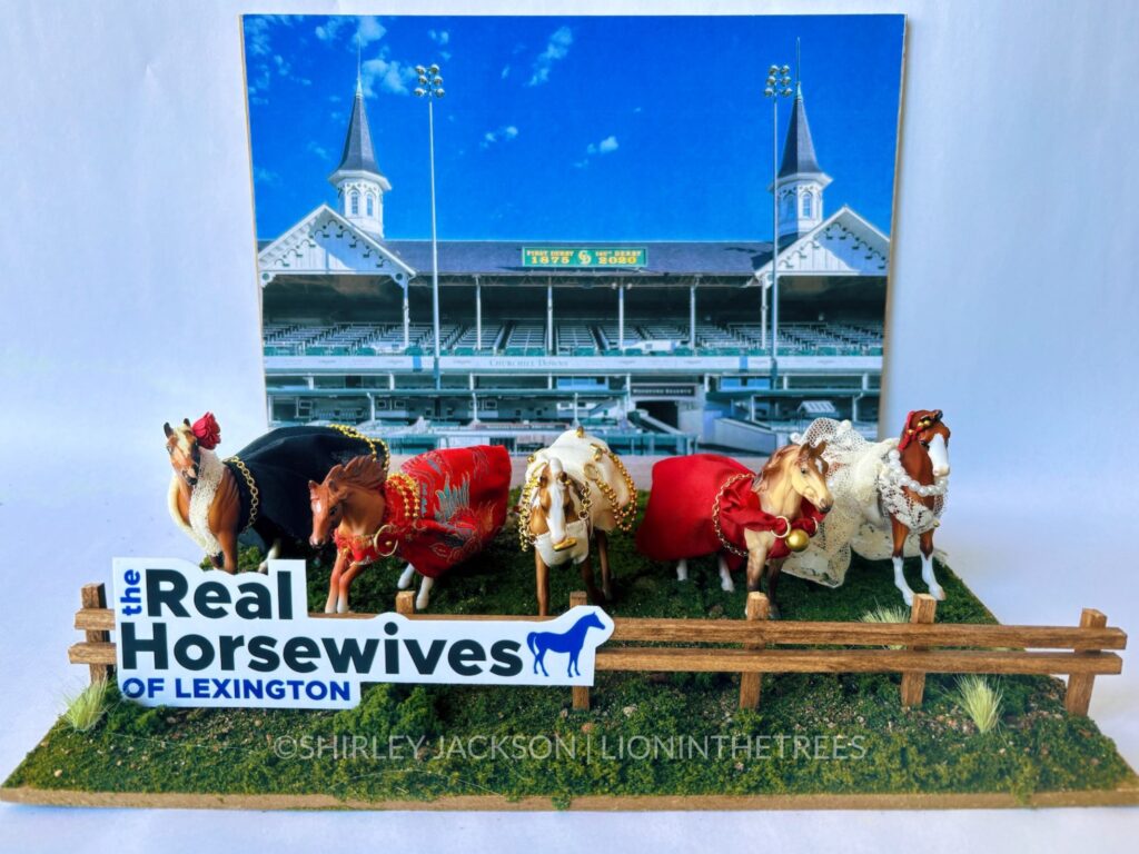 Photo of my completed diorama. There is a photo of Churchill Downs behind 5 horses dressed in different handmade clothing that are positioned on a grassy base with a brown fence in front of them. On the fence is "The Real Horsewive of Lexington" logo done in a very similar way as the actual logos found in the Real Housewives franchise.