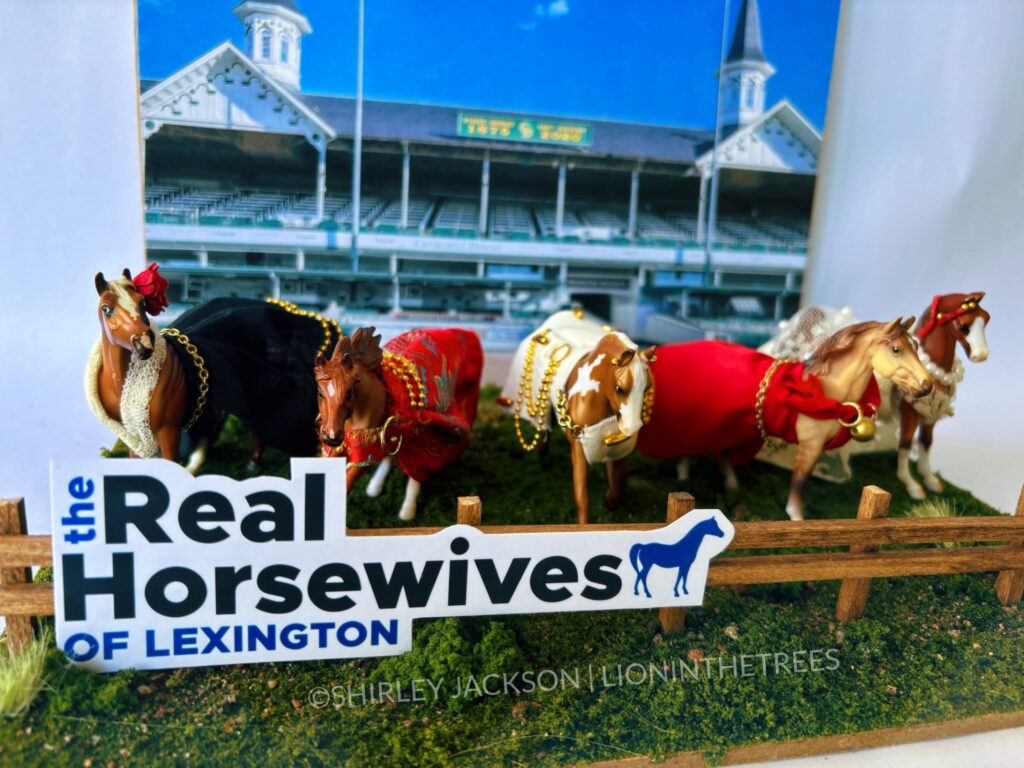 Close up photo of my completed diorama. There is a photo of Churchill Downs behind 5 horses dressed in different handmade clothing that are positioned on a grassy base with a brown fence in front of them. On the fence is "The Real Horsewive of Lexington" logo done in a very similar way as the actual logos found in the Real Housewives franchise.