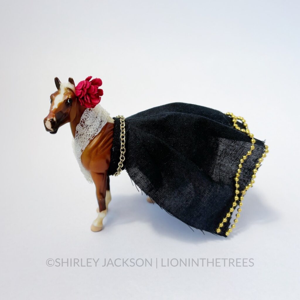 A chestnut horse dressed with a lace top and a black skirt with gold beads. She has a flower in her hair.