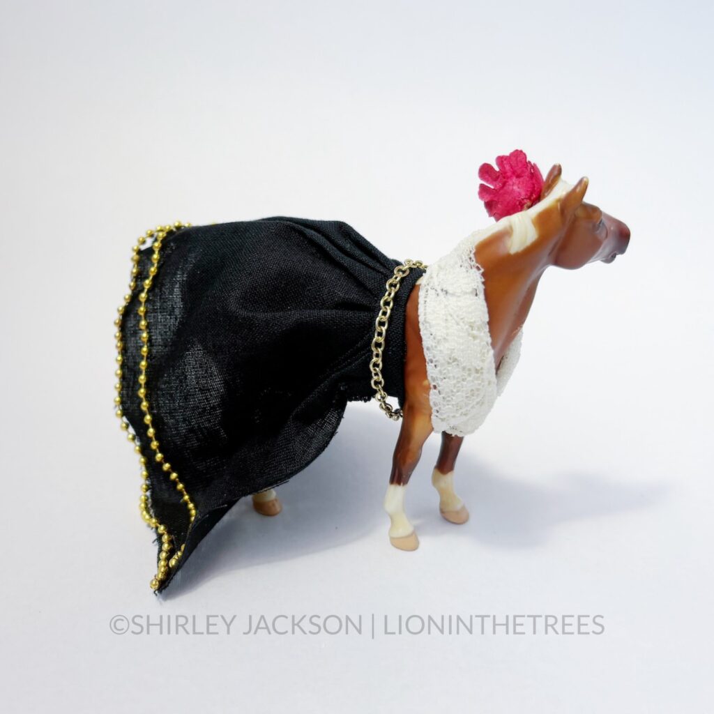 A chestnut horse dressed with a lace top and a black skirt with gold beads. She has a flower in her hair.