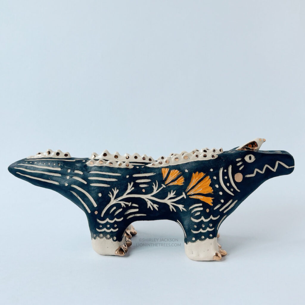 Right side view of the large alligator totem. This shows the carved California Poppy on it's side. They also have a little gold and white bird perched atop their head. This piece was done with black and orange underglaze, and gold overglaze details.