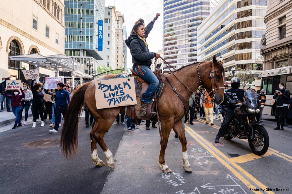 Photo of Brianna Noble on top of Dapper Dan in the center of a street in Oakland during the BLM protests of 2020 in the wake of the murder of George Floyd.