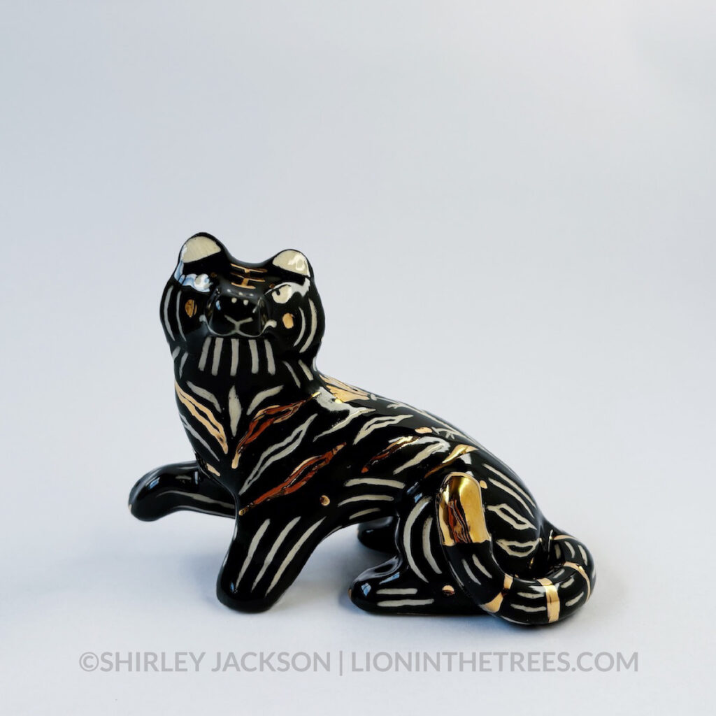 Front view. Sgraffito Black Tiger totem done with black underglaze and gold overglaze for some of the stripe details. It has one paw up in mid-air.