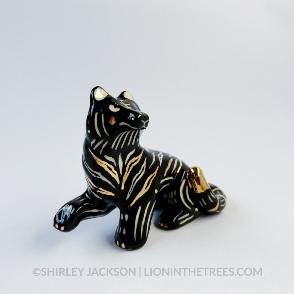 Front 3/4 view. Sgraffito Black Tiger totem done with black underglaze and gold overglaze for some of the stripe details. It has one paw up in mid-air.