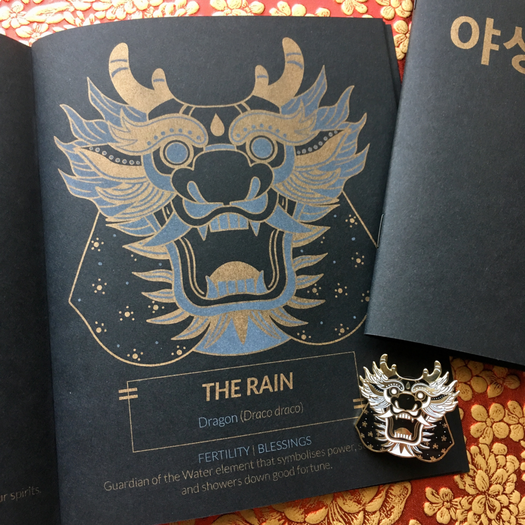 Close-up of my Oracles of the Wild risograph zine. This zine was printed with metallic gold ink and white ink on black paper. This photo portrays the Oracle The Rain, a mask based off of the Dragon.