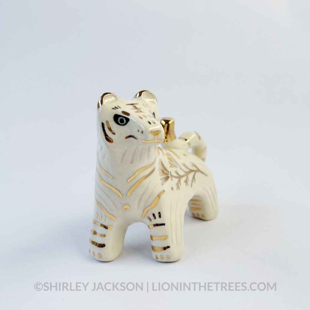 Front view of a sgraffito white tiger ceramic sculpture with gold overglaze details that make up the stripes, other markings, and my signature California Poppy motif on the side of it's body.