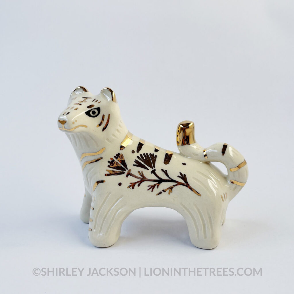 Left side view of a sgraffito white tiger ceramic sculpture with gold overglaze details that make up the stripes, other markings, and my signature California Poppy motif on the side of it's body.