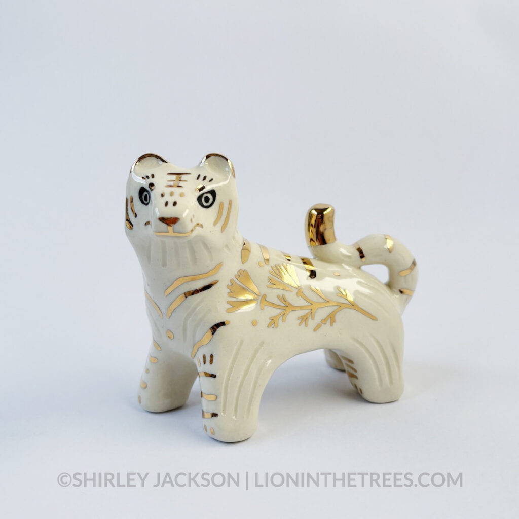 Front view of a sgraffito white tiger ceramic sculpture with gold overglaze details that make up the stripes, other markings, and my signature California Poppy motif on the side of it's body.