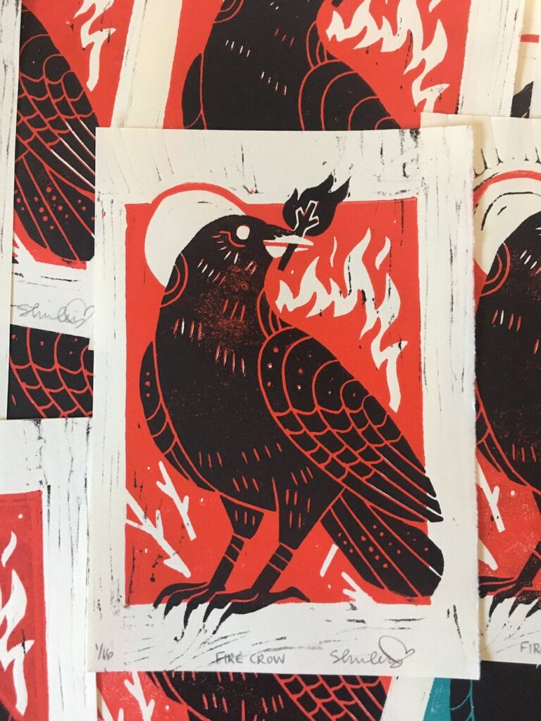 Linoblock relief print featuring a crow with a stick on fire in it's mouth. This print was done with a red background colour.