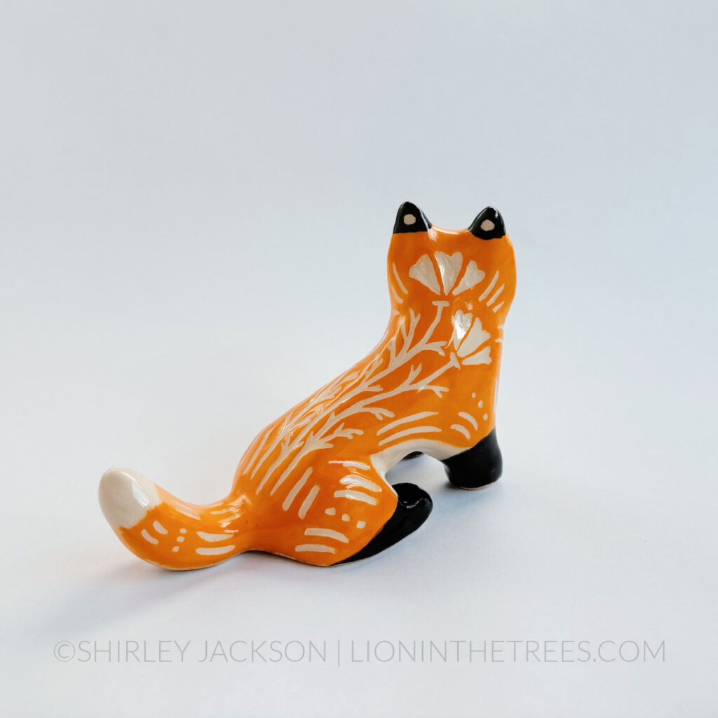 Sgraffito fox totem done with orange and black underglazes that mimics that colour pattern of a real fox. Finished with a clear glaze and carved California Poppies and various markings all over it's body. This is a back-side view of the piece.