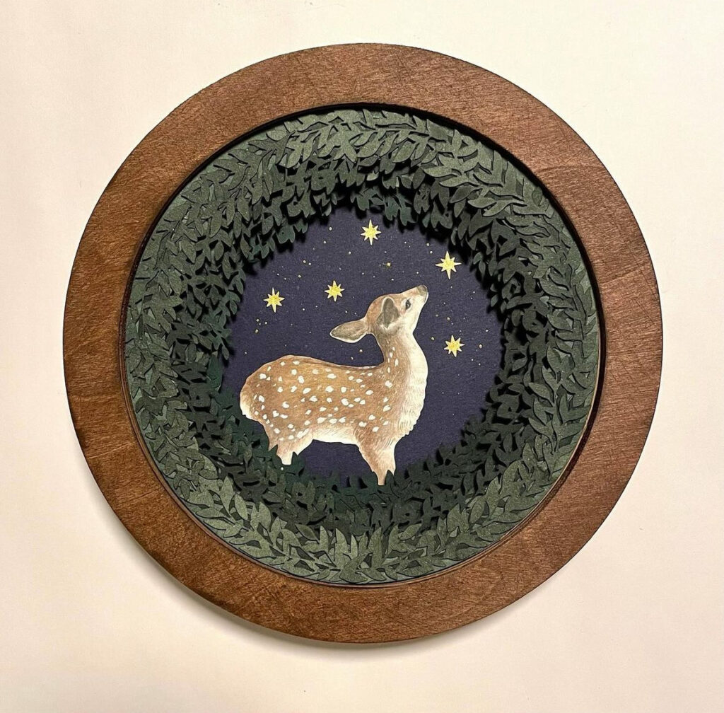 A circular wooden, shadow box that has these intricately carved flora that follows the circular wooden frame done in a green ombre the deeper you go into the piece. In the center is a fawn painted against a dark background and gold stars dancing around it.