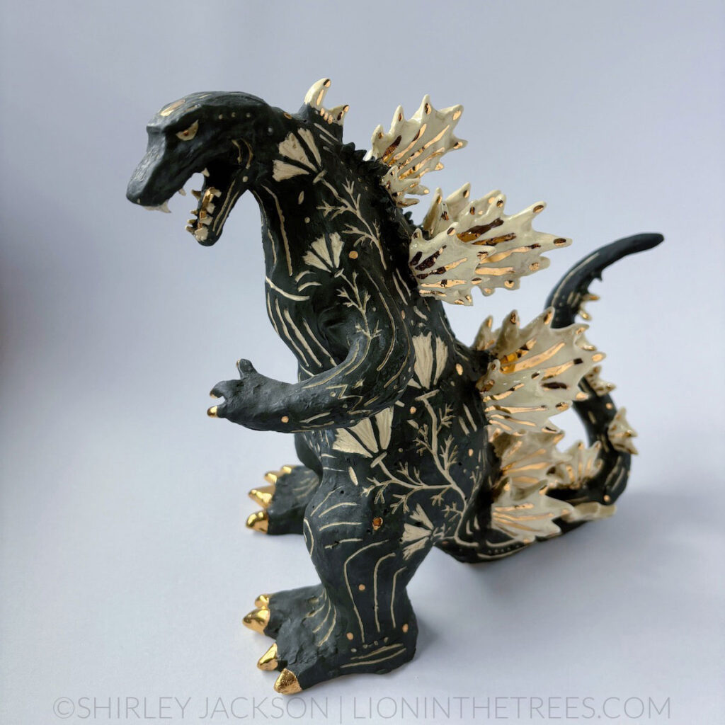 A black sgraffito Godzilla sculpture with California Poppy motifs carved all around it's body. There are gold overglaze details on his spines, claws, and toes.