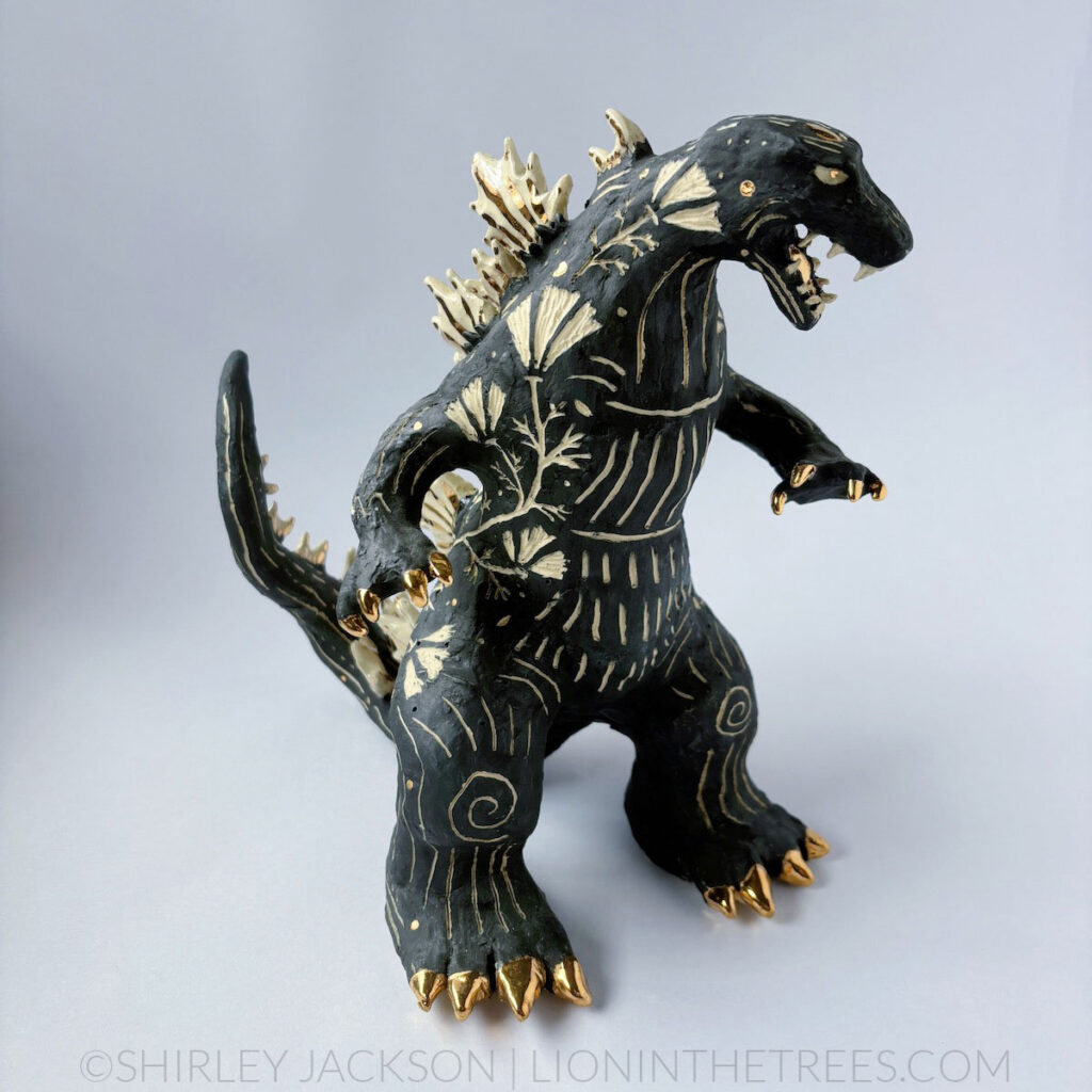 A black sgraffito Godzilla sculpture with California Poppy motifs carved all around it's body. There are gold overglaze details on his spines, claws, and toes.