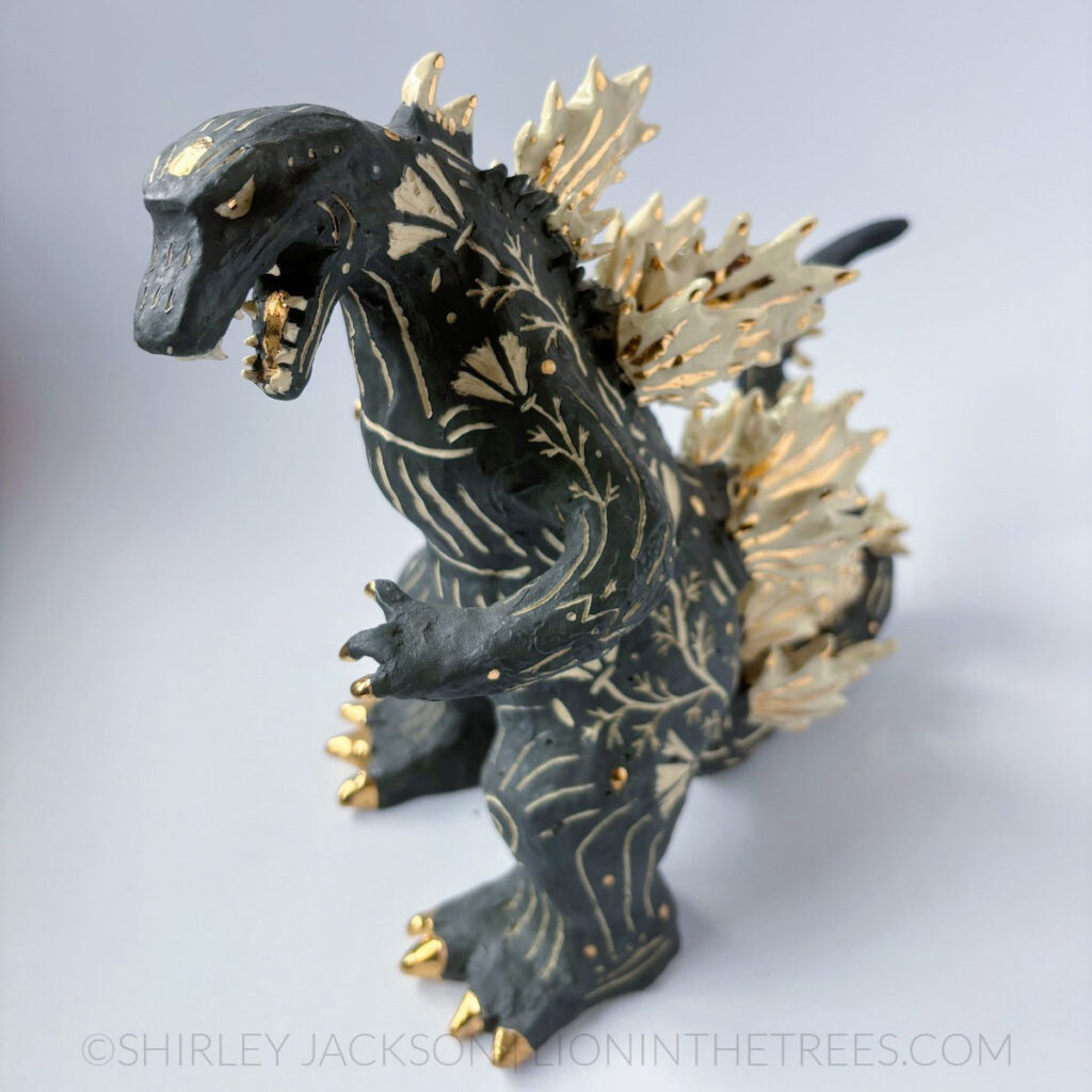 A black sgraffito Godzilla sculpture with California Poppy motifs carved all around it's body. There are gold overglaze details on his spines, claws, and toes.