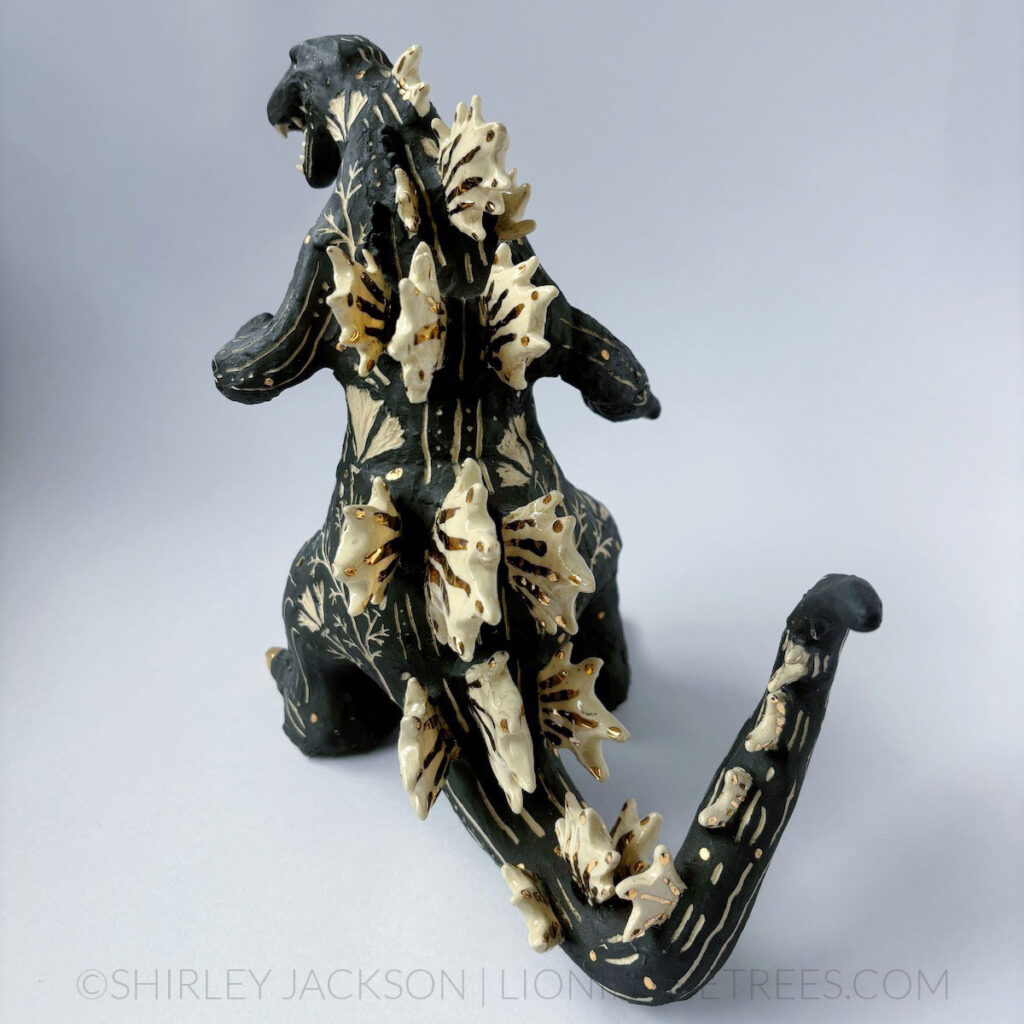 A black sgraffito Godzilla sculpture with California Poppy motifs carved all around it's body. There are gold overglaze details on his spines, claws, and toes.